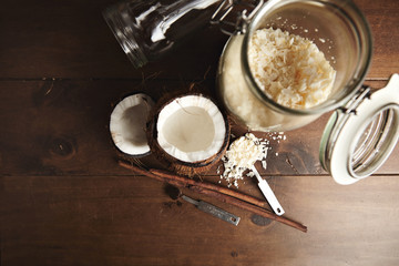 Opened fresh coconut next totransparent jar with coconut flakes, cinnamon stick and vintage steel measure spoons with natural brown sugar or grinded coffee, isolated on right side of wooden table, top