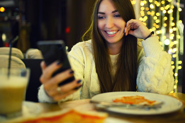 Woman typing write message on smart phone in a modern cafe. Cropped image of young pretty girl sitting at a table with pizza using mobile phone. Tonned. Selective focus.
