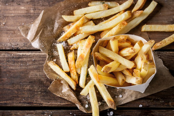 Fast food french fries potatoes with skin served with salt and herbs in lunch box on baking paper over old dark wooden background. Top view, space for text - obrazy, fototapety, plakaty
