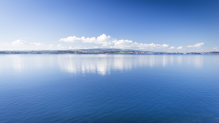 Clear day.  Lake Zug in central Switzerland. Clouds are reflected in water lake. At the edge water is clear and see the bottom of the lake.