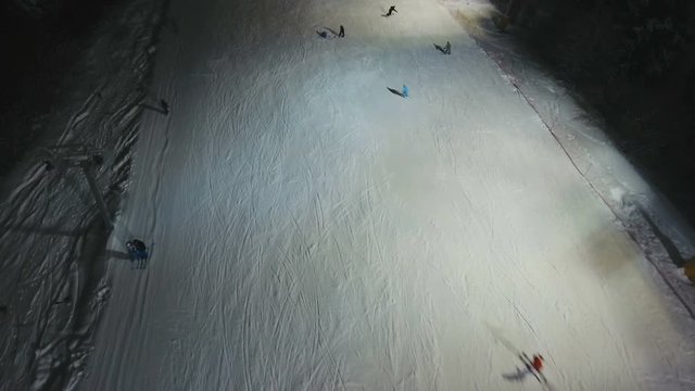 Aerial view: Skiers and snowboarders going down the slope in winter night. Skiers and snowboarders enjoying on slopes of ski resort in winter season. 4K video, aerial footage.