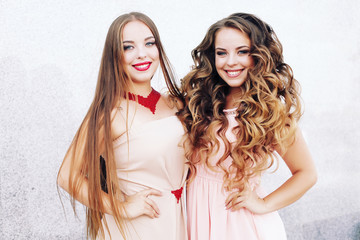 Two best friends embracing over gray wall. Young women having great time together. Sisters at the prom with gorgeous long curly hair. Professional make-up and hairstyle.      © perminoffa