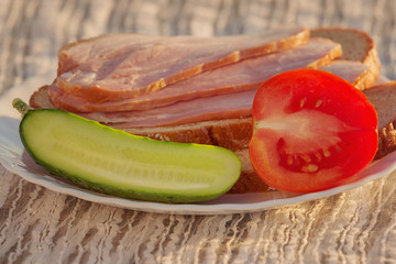 The plate is sliced ham, half a cucumber and half a tomato. Food closeup outdoors. Simple food, snack. Selective focus.
