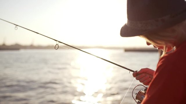 Young Caucasian kid in hat and red t-shirt checking his fishing rod near sea while sun shining in slowmotion