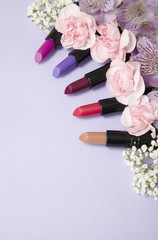 Obraz na płótnie Canvas Assorted colored lipstick make up and flowers arranged on a pastel purple background with empty space below