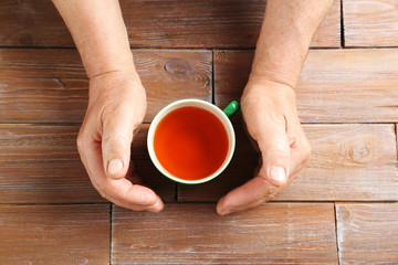 Hands of old senior with cup of tea on a wooden table