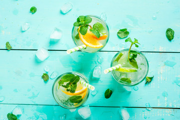 Selection of summer refreshing cocktails with mint (lemonade or mojito): with lemon, orange, lime. On wooden tray on light blue wooden table, bright sunlight. Copy space, top view