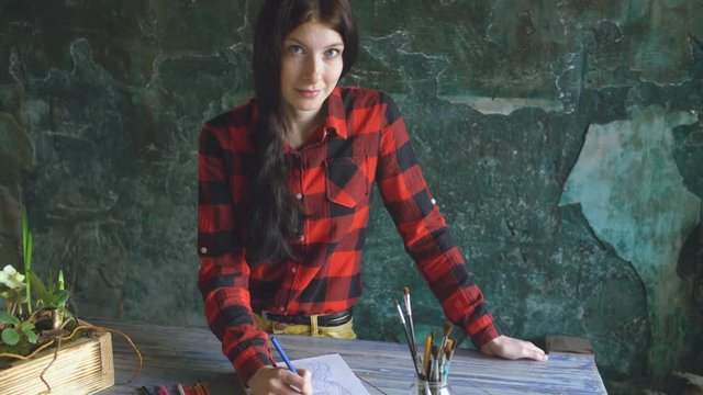 Young woman artist painting scetch on paper notebook looking into camera