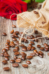 Roasted coffee beans, fresh red rose, coarse burlap sac on old wooden table. Vintage still life. Place for text. Top view.