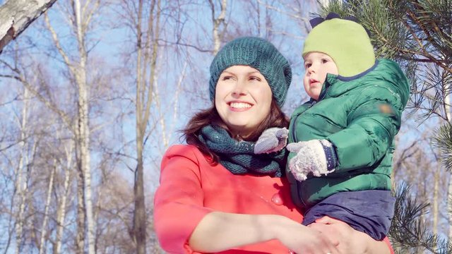 A woman in a red coat and green hat in the winter with a baby in her arms in Sunny weather, looking forward. Happy young family.