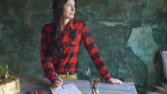Young woman artist painting scetch on paper notebook with pencil looks in window