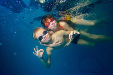 Fototapeta na wymiar Happy little boy and girl swim underwater, smiling, hugging and looking at the camera. Shooting under water. Portrait. Landscape orientation