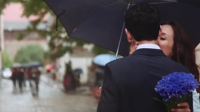 Few shots of beautiful young couple under the umbrella, attractive girl is hugging and kissing her boyfriend. Girl embraces her boyfriend. Couple goals. City settings on the background.
