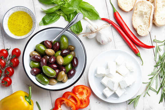 Mediterranean cuisine ingredients: fresh olives mix, rosemary twigs, green basil leaves, cherry tomatoes, pepper, feta cheese, ciabatta bread and olive oil on white wooden background
