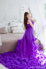 Fototapeta premium Beautiful woman in a long purple dress on a background of a large bed