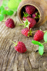 Red fresh raspberries on rough rustic wooden background. Cup with natural ripe organic berries with peduncles and raw green mint on table. Vertical, close up.