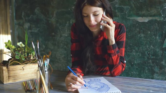 Young woman artist painting scetch on paper notebook with pencil. Girl smile and talks phone