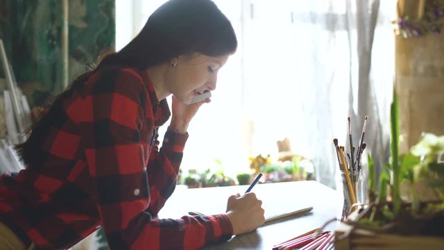 Young woman artist painting scetch on paper notebook with pencil. Girl smile and talks phone