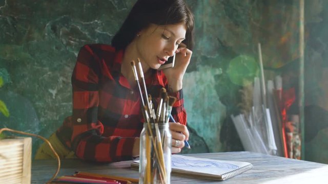 Young woman artist painting scetch on paper notebook with pencil. Girl smile and talks phone