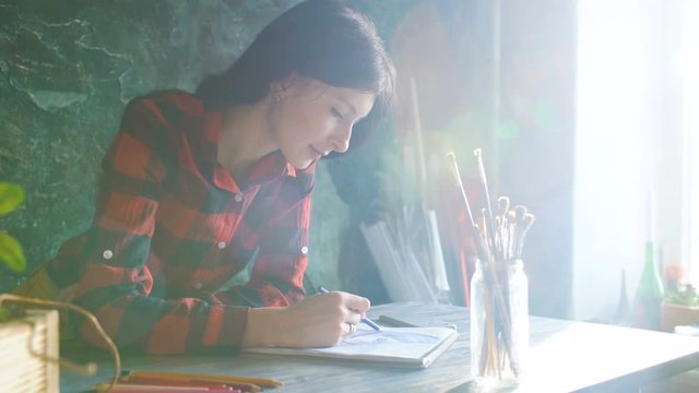 Young woman artist painting scetch on paper notebook with pencil. Bright sun flare from window