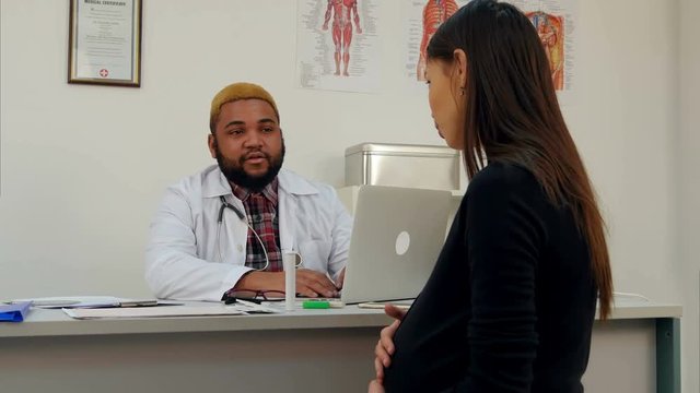 Pregnant woman visiting young smiling Afroamerican male doctor