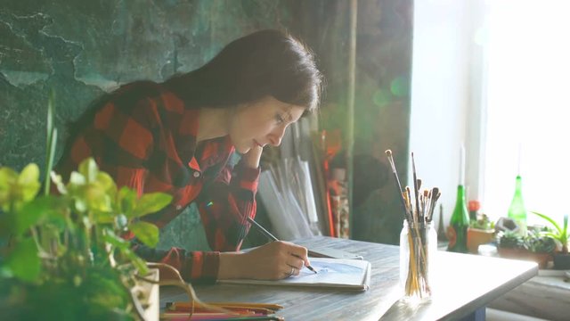 Young woman artist painting scetch on paper notebook with pencil. Bright sun flare from window