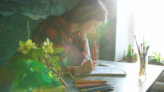 Young woman artist painting scetch on paper notebook with pencil. Bright sun flare from window