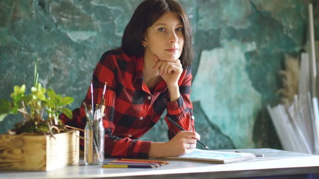 Young woman artist painting scetch on paper notebook with pencil and looks into camera smiling