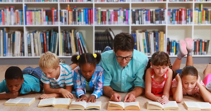 Teacher and kids lying on floor reading book in library at elementary school 4k