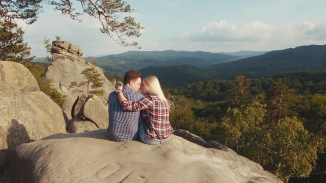 Attractive blonde girl and handsome young guy in a checkered stylish shirts. Lovely young couple is having a date on the top of a high rocky hill surrounded by forest. Romantic adventure. Sweet kisses