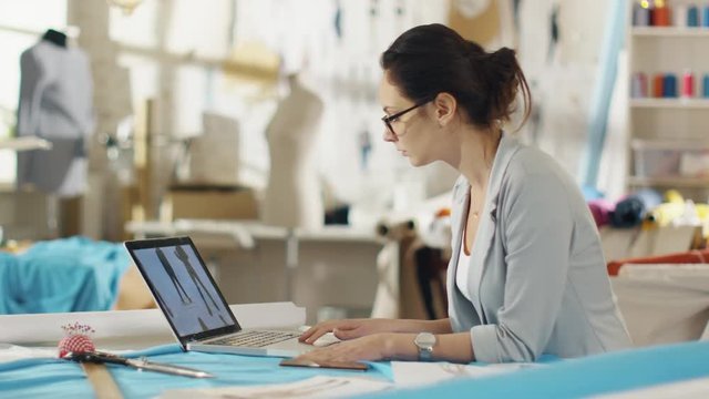 Beautiful Woman Fashion Designer Sitting at Her Table Working on Laptop, Looking at Drawings and Sketches