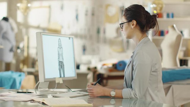 Beautiful Fashion Designer Sits at Her Desk before Personal Computer, Using Digital Drawing Tablet. Her Studio is Sunny, Full of Colorful Details, Fabrics, Sewing Items, Sketches. 