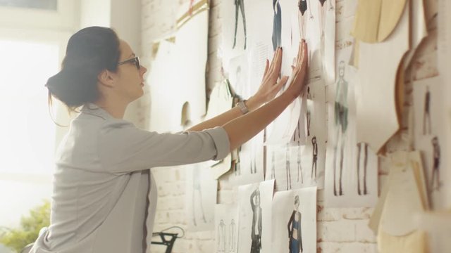 Female Fashion Designer Choosing Sketches for Her Project and Pinning them to the Wall. Her Studio is Sunny, Sewing Machine Stands on the Table.  Shot on RED EPIC 4K (UHD).