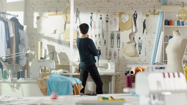 Female Fashion Designer Looking at Drawings and Sketches that are Pinned to the Wall. Studio is Sunny. Colorful Fabrics, Clothes Hanging and Sewing Items are Visible. Shot on RED EPIC 4K (UHD).