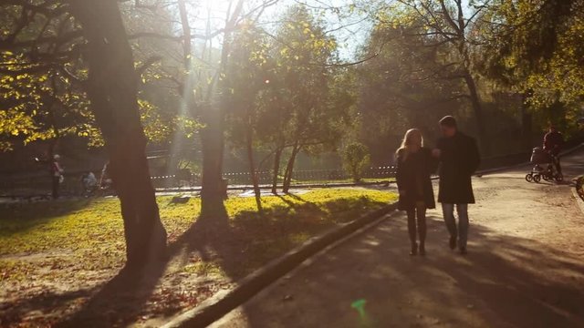 Amazing woman in a yellow dress and black coat, with a handsome man in elegant autumn wear. Luxurious young couple walking down the park alley and holding their hands. Being happy of being together.