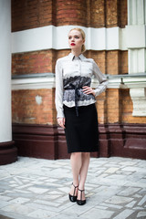 Obraz premium Happy Fashion blonde european elegant Woman with red lips and white skin Standing at the Old Red Brick building on green grass. Beautiful Female in formal black and white dress. 