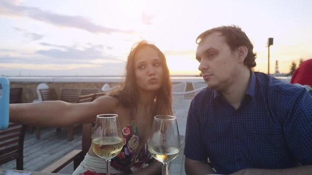 Young beautiful happy couple in love takes selfie with smartphone in outdoor cafe drinking wine at sunset time in slowmotion. 1920x1080
