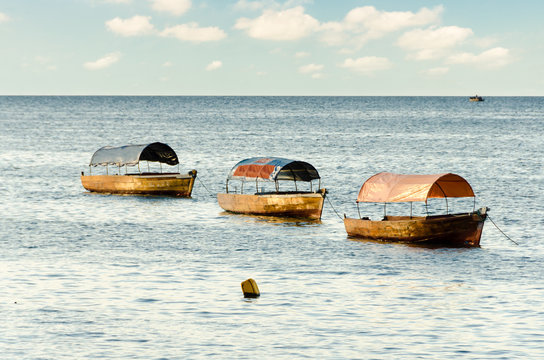 Three boats in line and tied to each other floating in the ocean off the coast of Zanzibar. It's a summer day with a few puffy clouds in the sky and calm sea.