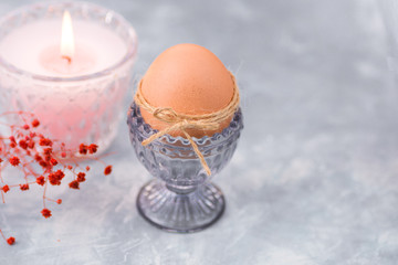 Easter brown egg in a crystal blueish cup, burning pink candle in the background,tender red psring flowers,blue and white concrete background,decoration concept