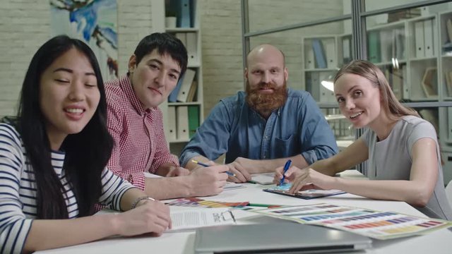 Group of young designers sitting at meeting table, smiling and waving at camera during web conference