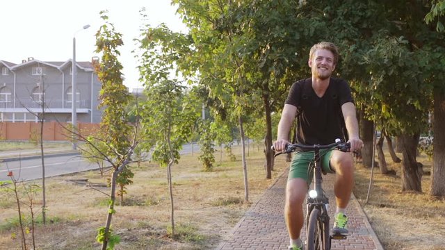 Handsome tall Caucasian male with light beard and black t-shirt riding bicycle in park smiling in slowmotion
