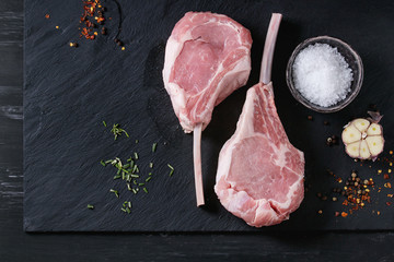 Two raw uncooked Veal tomahawk steak with garlic, salt and seasoning on black stone slate board. Top view with space.