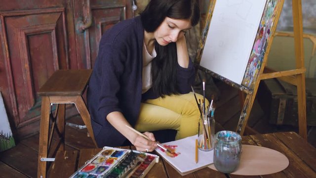 Young woman artist draw pictrure with watercolor paints and brush on easel canvas