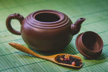 Close-up ceramic teapot, aromatic dry tea with fruits and petals in wooden spoon on  green bamboo...