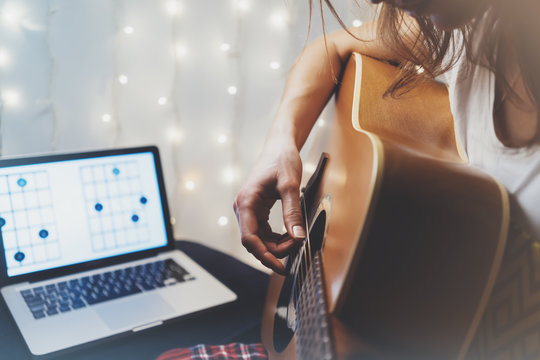 Hipster playing guitar in home atmosphere, person studying on musical instrument, notes in laptop on background glow bokeh illumination, female hands in holiday on relax glitter