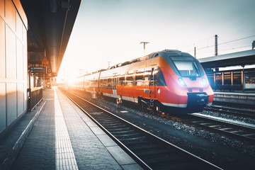 Obraz premium Nowoczesny szybki czerwony pociąg podmiejski na stacji kolejowej o zachodzie słońca. Włączam reflektory pociągu. Linia kolejowa z tonowaniem vintage. Pociąg na peronie. Krajobraz przemysłowy. Turystyka kolejowa