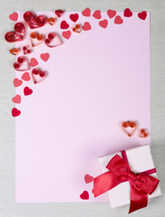 Pink paper sheet surrounded by handmade red and orange paper hearts and small square gift box with satin bow. Vertical orientation. Can be used as a Valentine's day or wedding frame or greeting card.