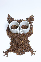 Roasted grains of black coffee. The grains are scattered in the form of an owl figurines. Owl Eyes in the form of two white cups and pour them coffee beans.