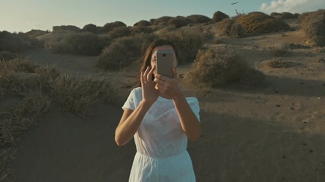 Girl in white dress makes the photo on the phone on a sunset, in desert. Warm summer evening, desert. Slow motion