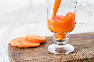 Carrot juice is poured into a glass Cup on a stem which stands on the table and the chopping Board,...